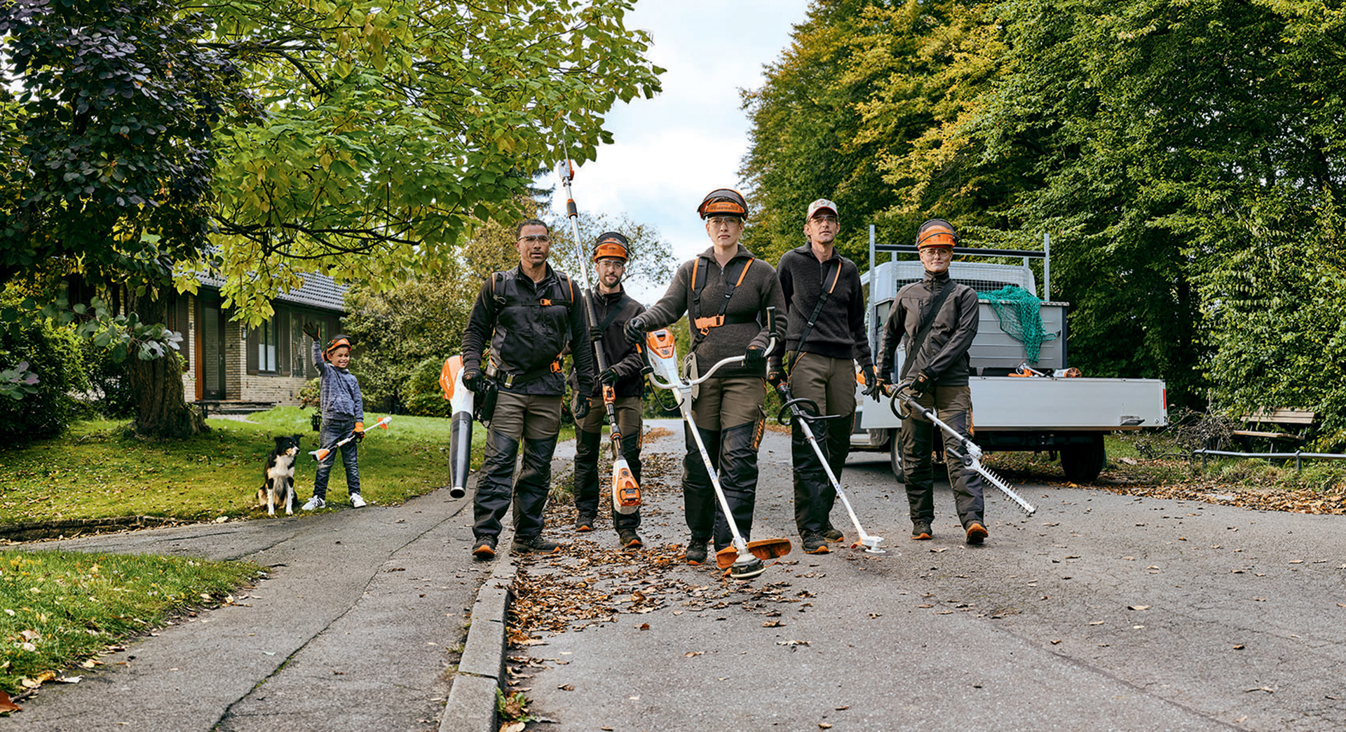 O echipă de horticultori și peisagiști cu utilaje cu acumulator profesionale STIHL se plimbă pe o stradă.